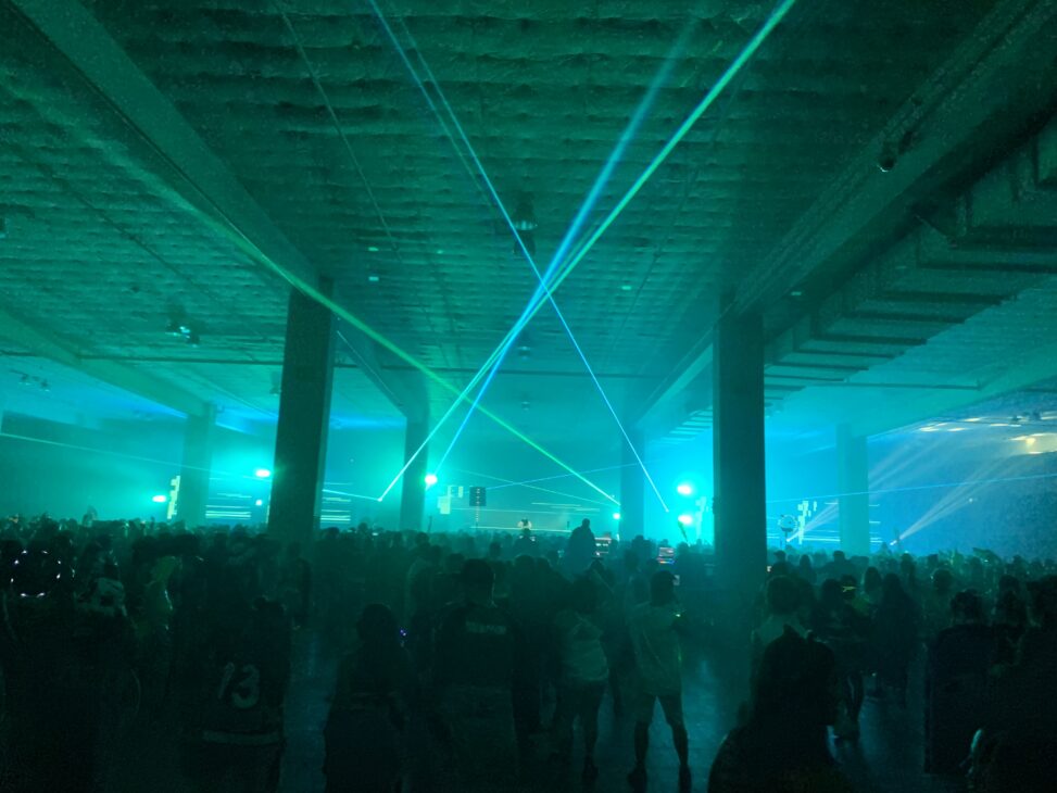 Lasers and strobe lights illuminate a dim parking garage, full of people dancing with a stage at the back of the room.
